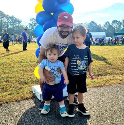 A white father stands with his two white sons, all smiling, in front of a blue and yellow balloon arch at an outdoor festival.