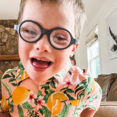 A close up of a white school aged boy with Down syndrome wearing glasses smiling at the camera.