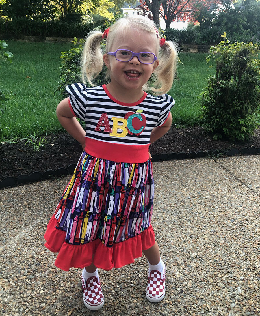 A young white girl with Down syndrome with blonde hair in pigtails wearing glasses and a dress with ABC on the front smiling at the camera.