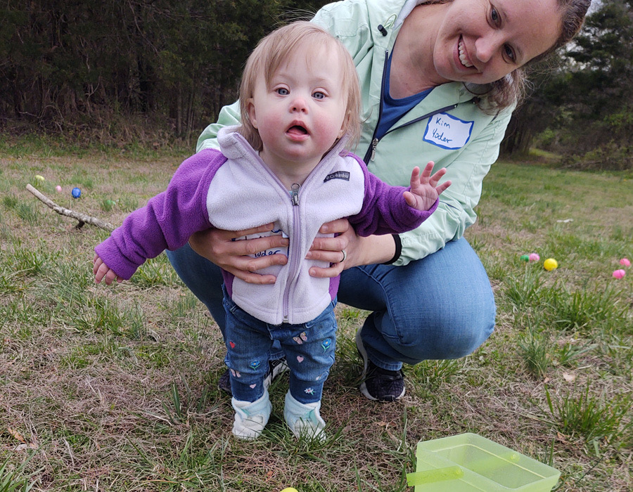 A white woman kneeling behind her white toddler daughter with Down syndrome, holding her by her hips, smiling.