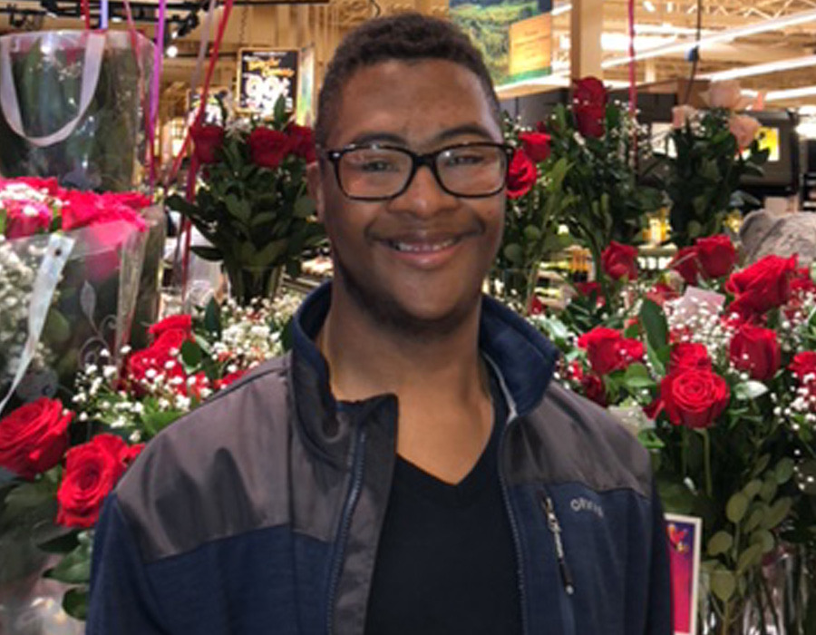 A young Black man with Down syndrome wearing glasses and a jacket, standing in front of a display of roses, smiling and looking at the camera.